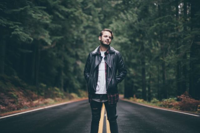 man-wearing-black-leather-jacket-standing-on-highway-in-the-middle-of-green-trees