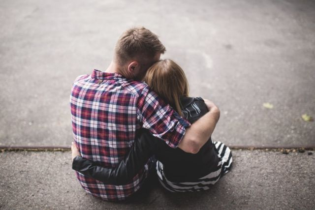 man-in-red-white-and-blue-check-long-sleeve-shirt-beside-woman-in-black-and-white-stripes-shirt-hugging-each-other-while-sitting-on-a-concrete-surface