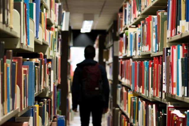 person-wearing-blue-black-jacket-standing-near-gray-bookshelf