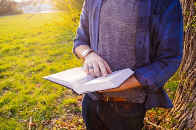 man-wearing-blue-and-black-flannel-shirt-holding-white-and-black-book-during-daytime