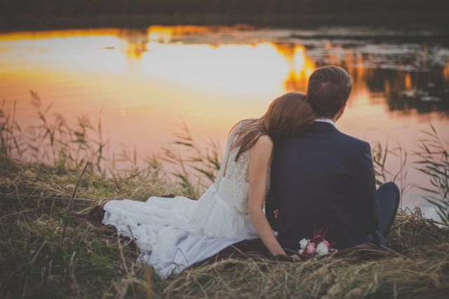 wedding-couple-sitting-on-green-grass-in-front-of-body-of-water-at-sunset